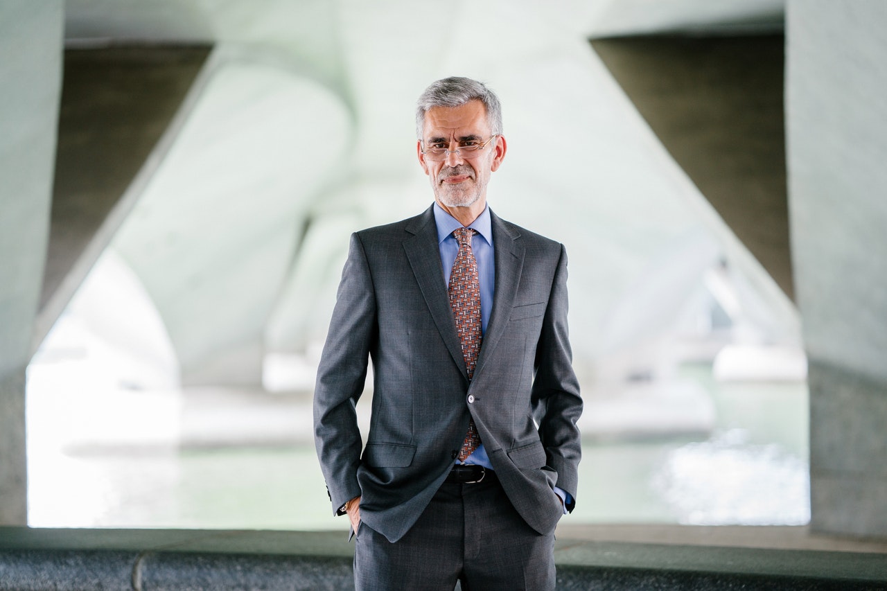 A confident man in a suit smiles slightly with his hands in his pockets.